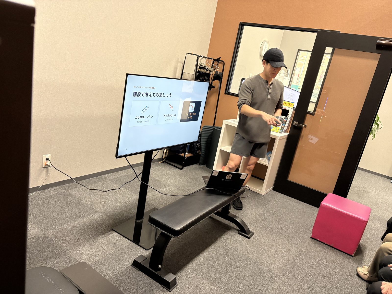 比嘉一雄さんが「階段で考えてみましょう」というスライドの横で説明している。スライドには「上るのは、つらい」「下りるのは、楽」「でも、効くのは下りる」とある。